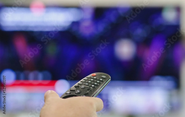 Fototapeta Close-up of hand with the remote control television and presses the button. Television remote control changes channels thumb on the blue TV screen.