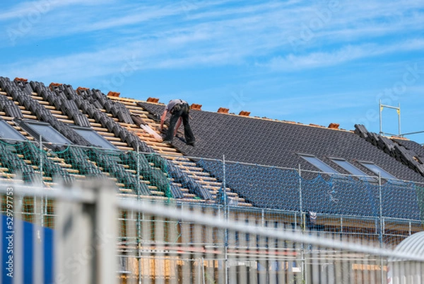 Fototapeta Dachdecker deckt das Schrägdach mit Holzgerüst eines Neubaus mit dunklen Dachziegeln bei blauem Himmel, selektiver Fokus, viel Copy space