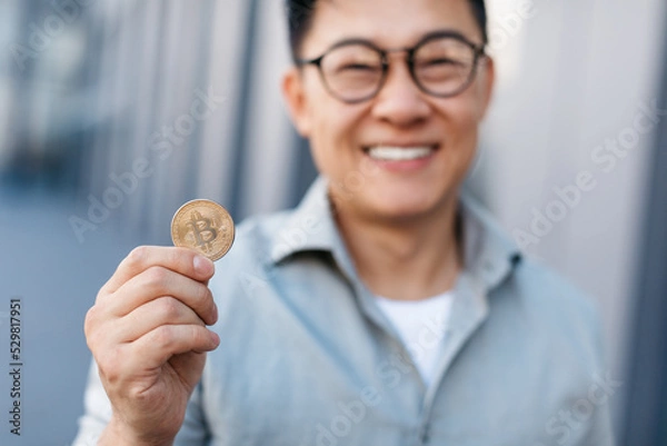 Fototapeta Smiling asian middle aged businessman showing bitcoin, standing near modern office building, selective focus