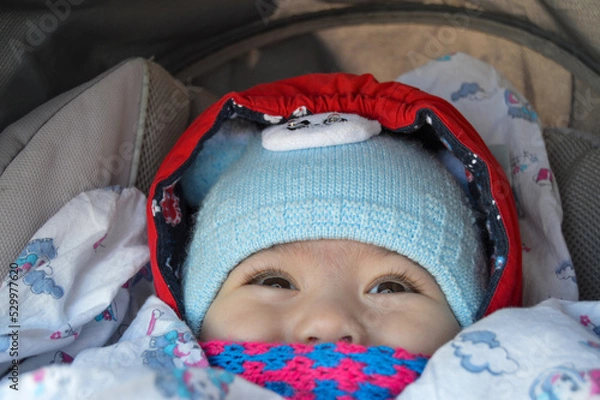 Obraz The child is sitting in a stroller with a pink scarf and a blue hat, only the eyes and nose are visible.
