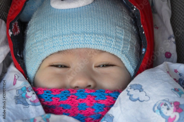 Obraz The child is sitting in a stroller with a pink scarf and a blue hat, only the eyes and nose are visible.