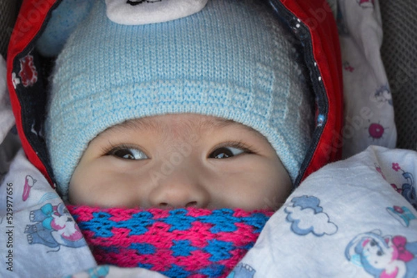 Obraz The child is sitting in a stroller with a pink scarf and a blue hat, only the eyes and nose are visible.