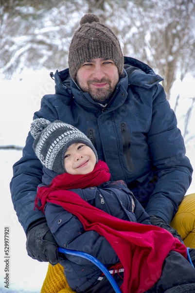 Obraz Father and son sledding in the snow in winter. Winter entertainment and recreation. The man and child are smiling into the frame. Warm clothes winter season