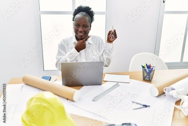 Obraz Young african woman working at architecture studio holding house keys smiling happy pointing with hand and finger