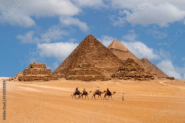 Fototapeta Cairo, Egypt. 08.25.2022. Group of tourists riding camels seeing the pyramids of Menkaure, Chephren and Cheops.