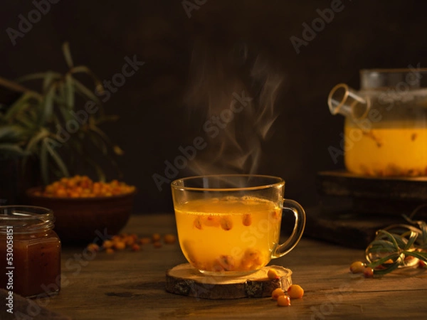 Fototapeta Sea buckthorn tea in a glass cup with steam. Dark background. Next to the cup is a teapot, a bowl of sea buckthorn berries and a jar of sea buckthorn jam.