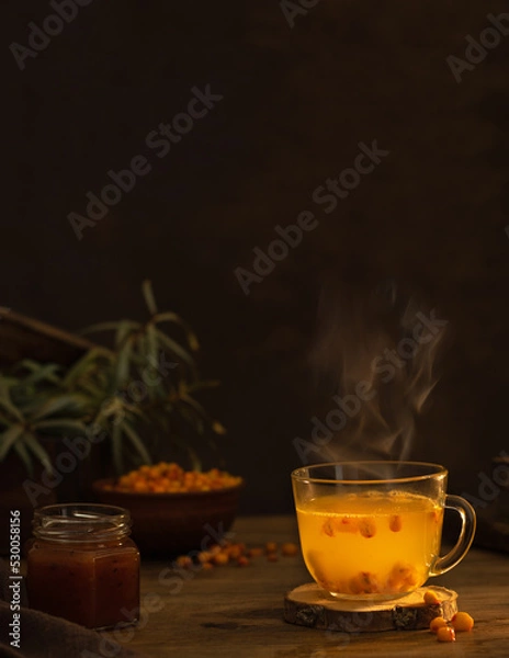 Fototapeta Sea buckthorn tea in a glass cup with steam. Dark background. Next to the cup is a bowl of sea buckthorn berries and a jar of sea buckthorn jam.