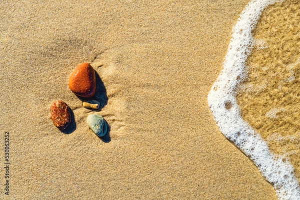 Fototapeta Varicolored stones on the sand with a wave and with foam. Top view, closeup. Background.