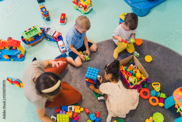 Fototapeta Toddlers and their nursery teacher playing with plastic building blocks and colorful car toys while sitting on the floor in a playroom. Early brain and skills development. High quality photo