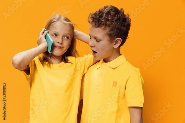 Fototapeta cute funny school-age children stand on a yellow background in bright clothes and hugging each other talking on the phone