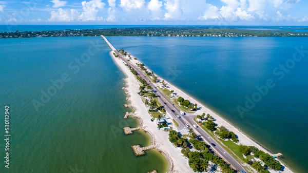 Fototapeta Aerial Drone Distanced View of the Causeway Bridge in Sanibel, Florida with the Bay and a Preserve in the Foreground and the Gulf of Mexico in the Background Featuring a Blue Sky and Blue Water