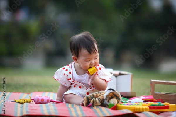 Fototapeta An Asian baby is sitting on a colorful mat eating a yellow block toy with other toys surrounding her in blur background.