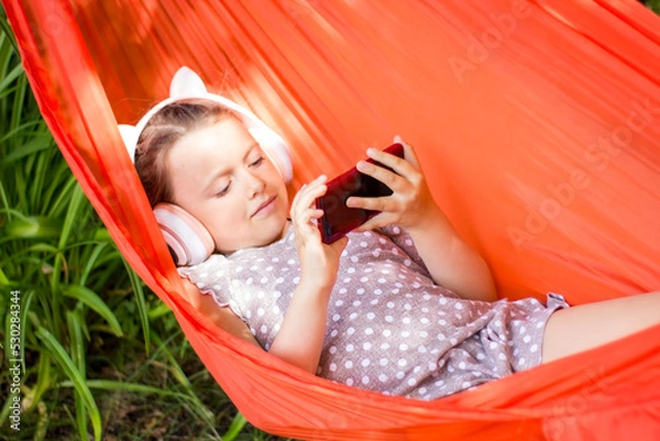 Obraz Cute little girl lies in a hammock with smartphone and wireless headphones and listening to music.