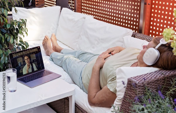 Fototapeta Adult senior plus size woman having online hypnotherapy session with psychotherapist lying on sofa on outdoor terrace at home.