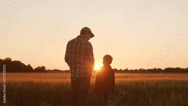 Obraz Farmer and his son in front of a sunset agricultural landscape. Man and a boy in a countryside field. Fatherhood, country life, farming and country lifestyle.