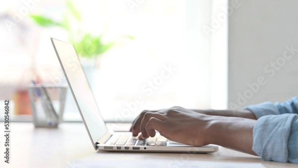 Fototapeta Side View of Young African Man Typing on Laptop Keyboard
