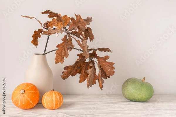 Fototapeta Autumn season concept. Dry oak leaves in vase with decorative pumpkins on light wooden table.