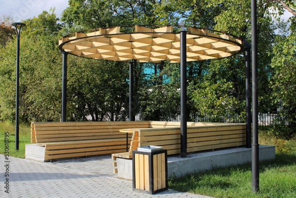 Fototapeta A light wooden gazebo with a bench, a table and an unusual hutch in a city park. Urban architecture