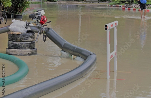 Fototapeta Pipes lead to a pump working on a flooded residential street. A flood barrier warns that the machine is in operation.  Blurred in the background a person can be partially seen walking in the water.