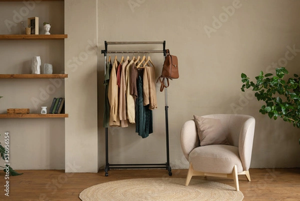 Fototapeta Rail rack with a capsule wardrobe in the living room against a beige wall. Trendy outfits in autumn colors: burgundy, beige, khaki, green, brown.