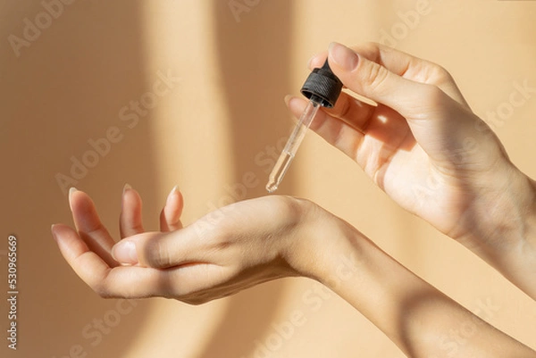 Obraz Women's hands hold pipette with cosmetic serum on light brown background, in rays of sunlight. Close-up, copy space. Concept of body care