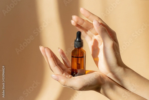 Obraz Women's hands hold bottle of amber glass with cosmetic serum on brown background. Mockup of container with dropper lid with collagen in rays of sunlight. Concept of skin care