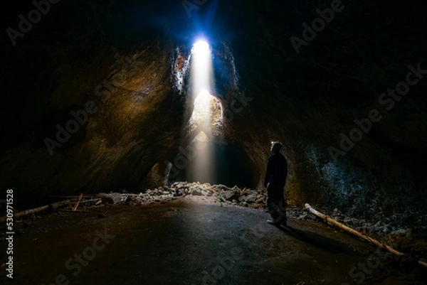 Fototapeta Adventurous athletic male standing in a lava tube looking at the sunlight shinning down into the cave through holes in the ceiling.