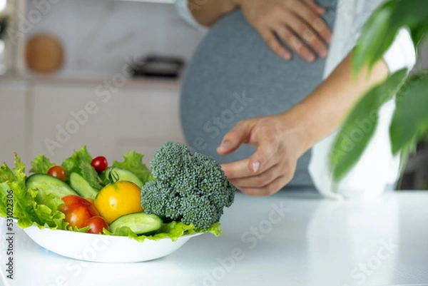 Obraz Pregnant woman stretch hand to plate with vegetables and fruits on table. Nutrition and healthy diet during pregnancy. Healthy eating full of vitamins and antioxidants for pregnant woman.