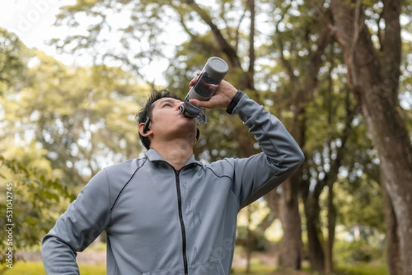 Fototapeta Thirsty runner man is drinking water bottle with earphone after exercise cardio on path in forest background.
