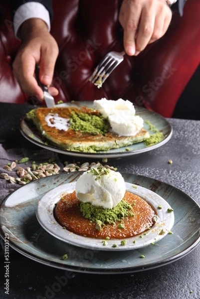Fototapeta Oriental sweets. Pahlava with ice cream nuts, pistachio in a large dish on a black table with hands. Turkish dessert. Background image, copy space, horizontal