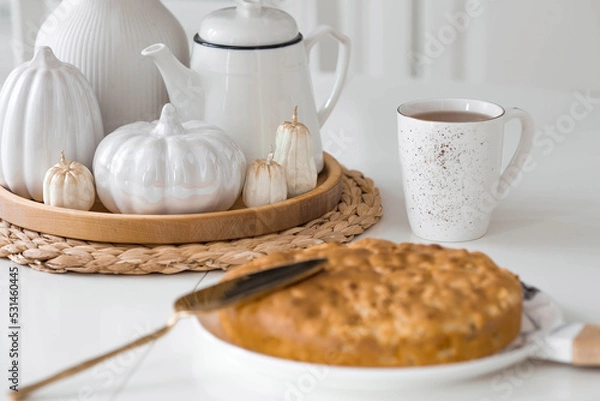 Fototapeta Still-life. Dried pampas grass in a vase, white ceramic pumpkins, candles and apple pie charlotte on a white table in the interior of a Scandinavian-style home kitchen. Cozy autumn concept.