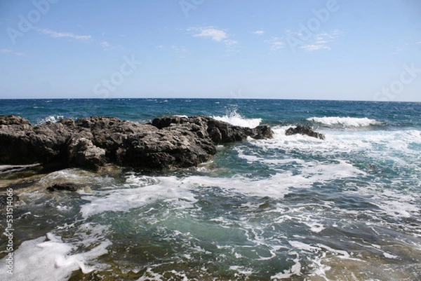 Fototapeta Waves crash over a rock in an aqua blue ocean with a blue sky on top.  Image has copy space.