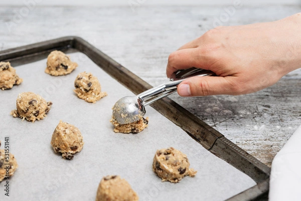 Fototapeta Hand of male baker scooping raw oatmeal raisin cookie dough with scoop on cookie sheet with baking paper, traditional and most popular American biscuit recipe made of flour, sugar, eggs, rolled oats