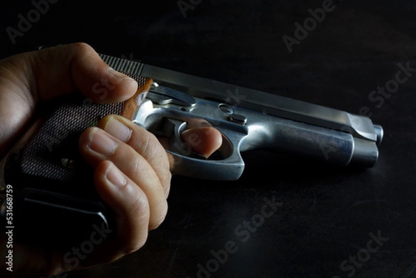 Obraz Left human hand holding gun. 9 mm parabellum semi automatic stainless steel pistol lay down on low light dark background. Low key pgotography.