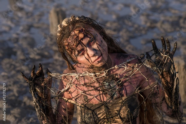 Fototapeta The concept of plastic pollution of the marine environment and conservation of nature - an injured mermaid tangled in a net, stained with fuel oil, suffering from dirt and debris.

