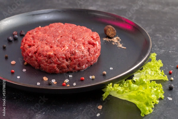 Obraz Chopped Steak With Nutmeg And Pappercorns On A Black Plate. The Hamburger Is Ready To Fry. Minced Beef. Fresh Lettuce Leaf As Garnish. Culinary Preparation. Side View. Close Up. Selective Focus.