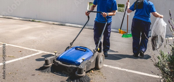 Fototapeta Two young latino janitorial workers (man and woman) in uniform cleaning in parking lot with outdoor industrial vacuum cleaner