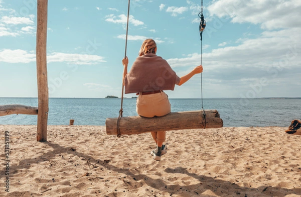 Obraz A beautiful woman is swinging on a swing overlooking the sea. Autumn at sea. Sand, water, swing. Rest by the water.