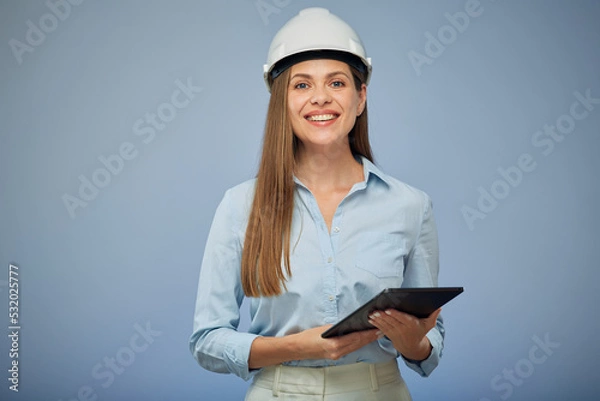 Obraz Smiling woman engineer in safety white helmet using digital tablet. Isolated female portrait on blue background.