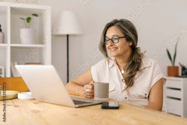 Fototapeta Woman holding a cup and reading from computer screen