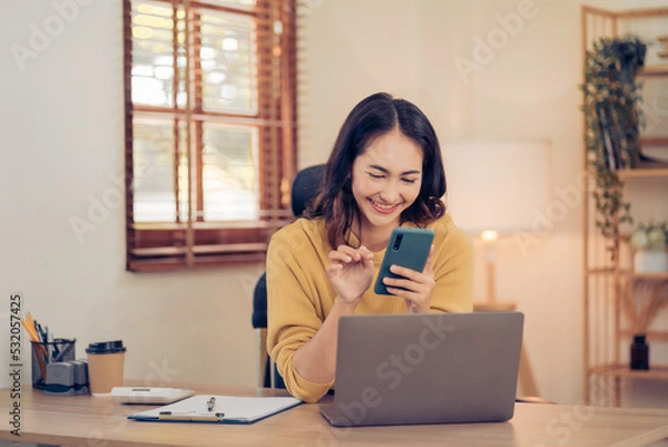 Fototapeta Attractive brunette Asian woman using mobile phone, smile and happy while sitting at working desk in modern office.