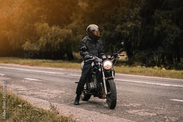Fototapeta male motorcyclist in a warm jacket and helmet in cold autumn weather on the road with a motorcycle cafe racer