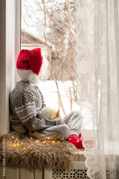 Fototapeta a child in festive clothes and a red Christmas hat is sitting by the window with a golden ball in his hand
