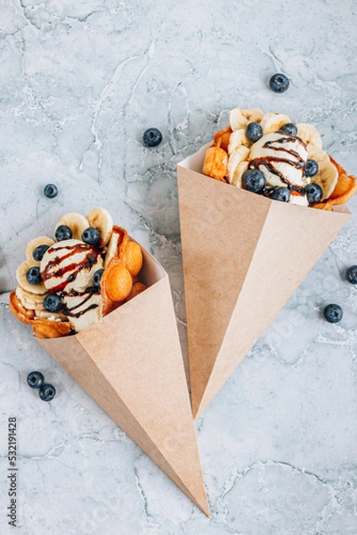 Fototapeta Bubble waffle with ice cream, banana and blueberry in paper cup over gray marble background