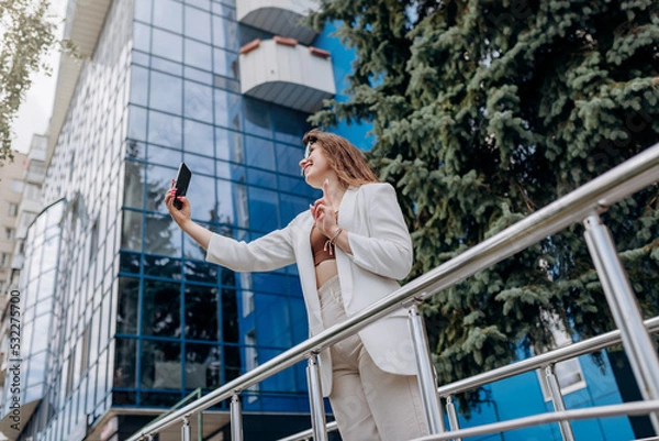 Fototapeta Smiling business woman in white suit and sunglasses making video call to friend using phone during break standing near modern office building