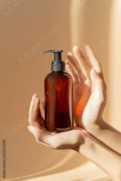 Obraz Women's hands hold bottle with dispenser for liquid soap, shampoo, lotion. Mock-up of an unmarked container in sunlight on beige background. Skin care concept, vertical image