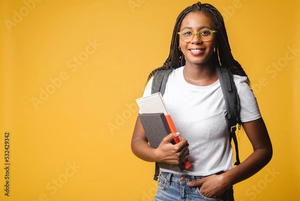 Obraz Inspired multiracial female student in white t-shirt and jeans with backpack isolated on yellow, foreigner woman in glasses proud to be university member, looking at the camera standing indoors