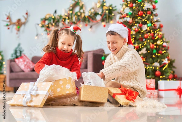Fototapeta cute cheerful children with an open smile, brother and sister, open golden gift boxes tied with red ribbon. children laugh and hold gifts in their hands. Christmas tree in light garlands in background