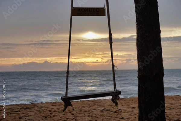 Fototapeta Close-up to a swing chair tied to a tree with a backdrop of the evening sea and the setting sun.