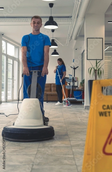 Fototapeta Senior cleaner cleans the hard floor with machine while cleaning lady cleans in the backround
